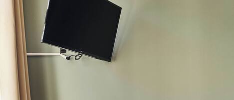 Una habitación con escritorio, televisión montada en la pared, cubo de basura y reposapiés de mimbre.