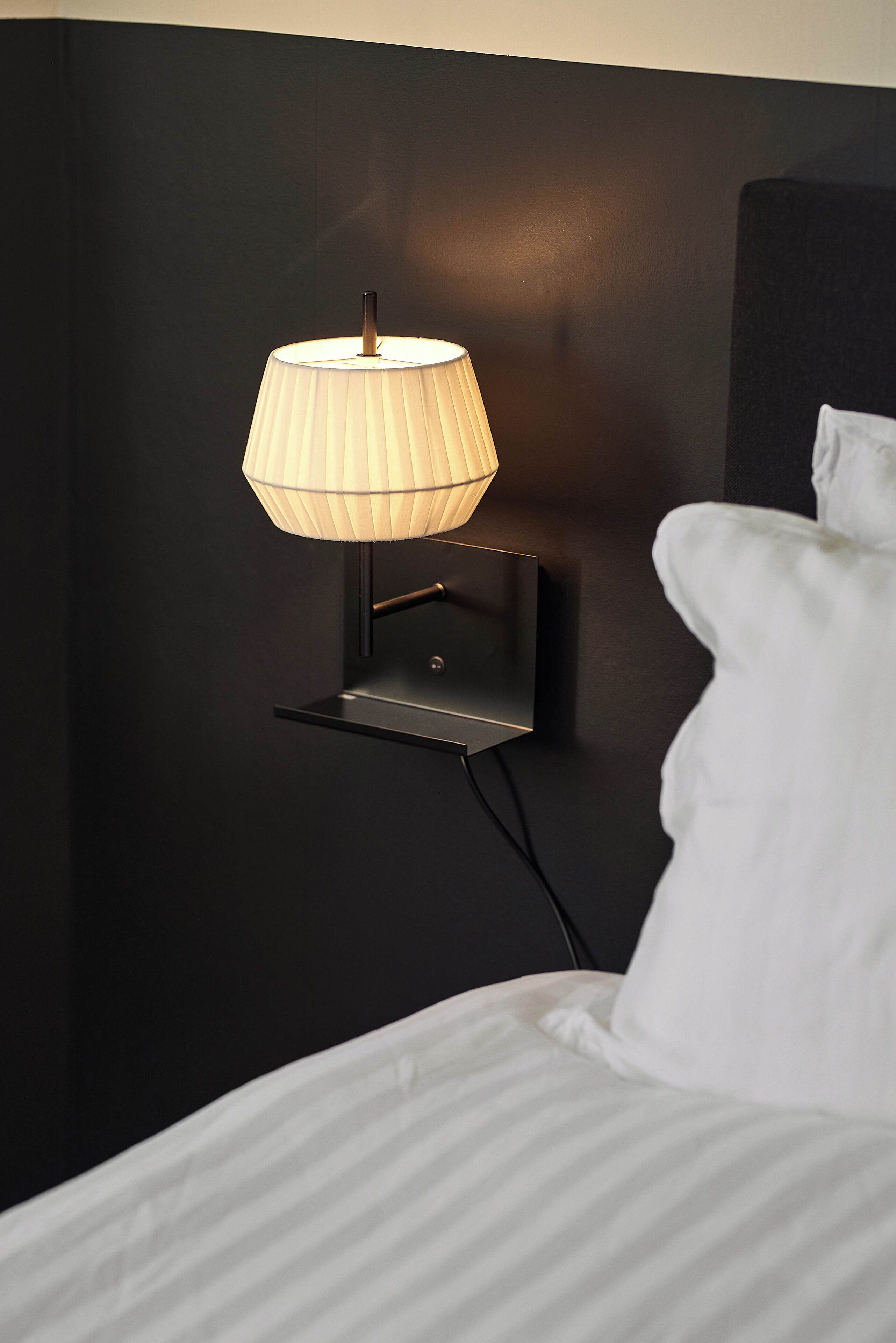 Una mesita de noche moderna con una lámpara de pared, pared negra y almohada blanca.