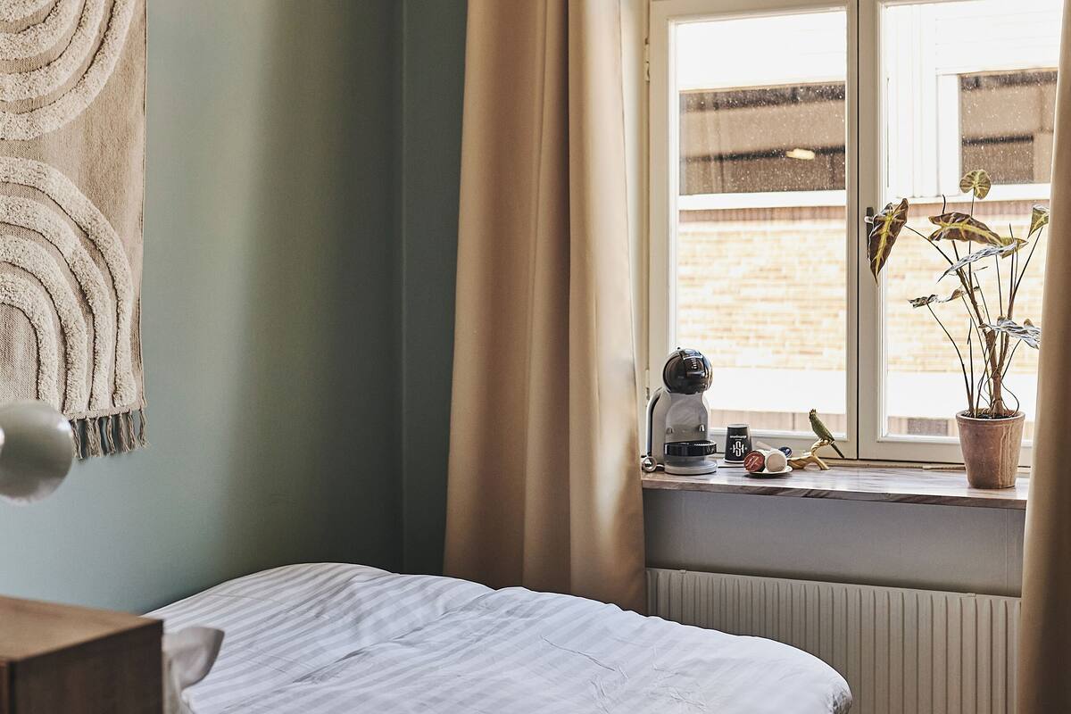 Un dormitorio con cama, ventana con cortinas, radiador y una planta en el alféizar.