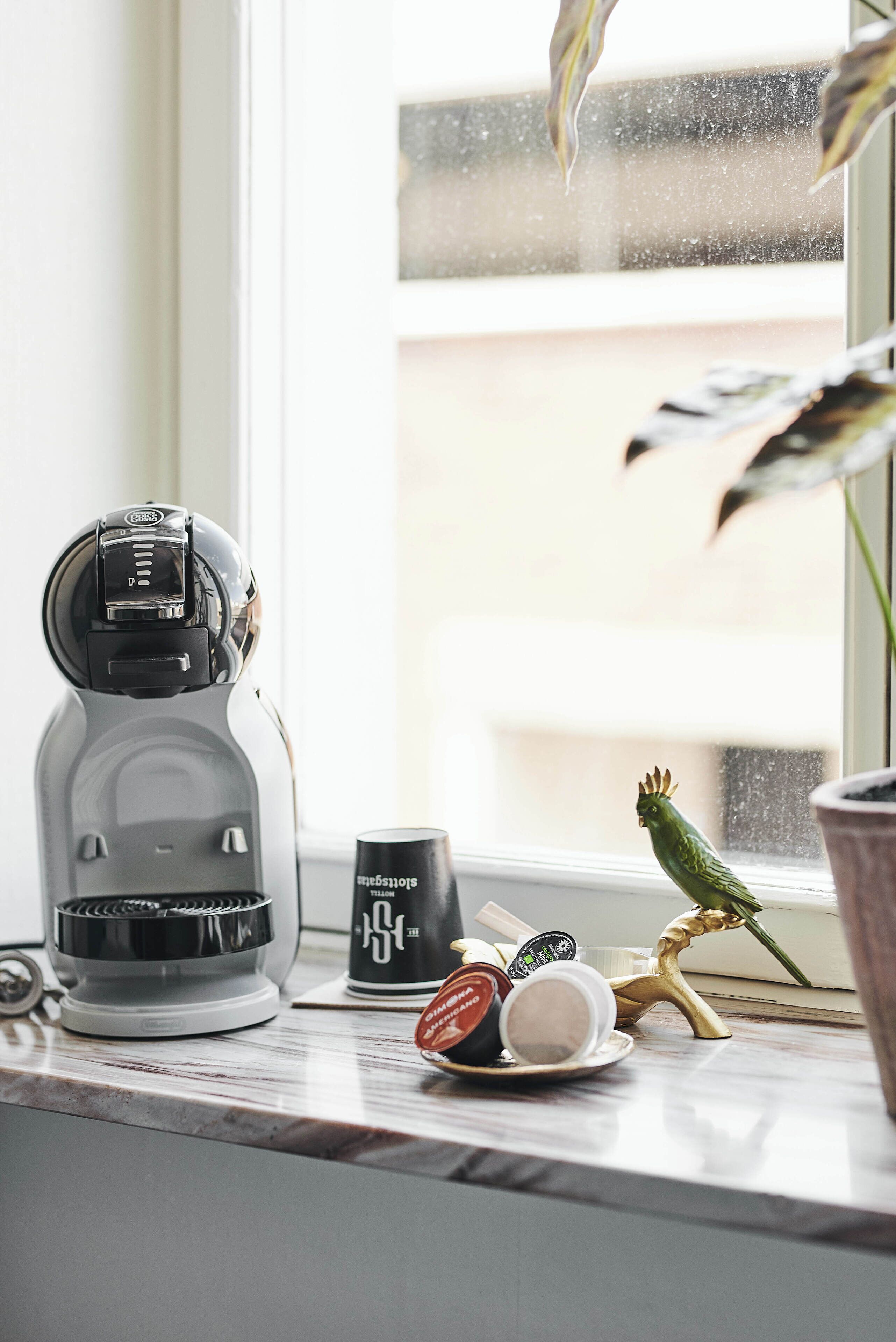 A coffee maker, a mug, and a bird figurine on a windowsill.