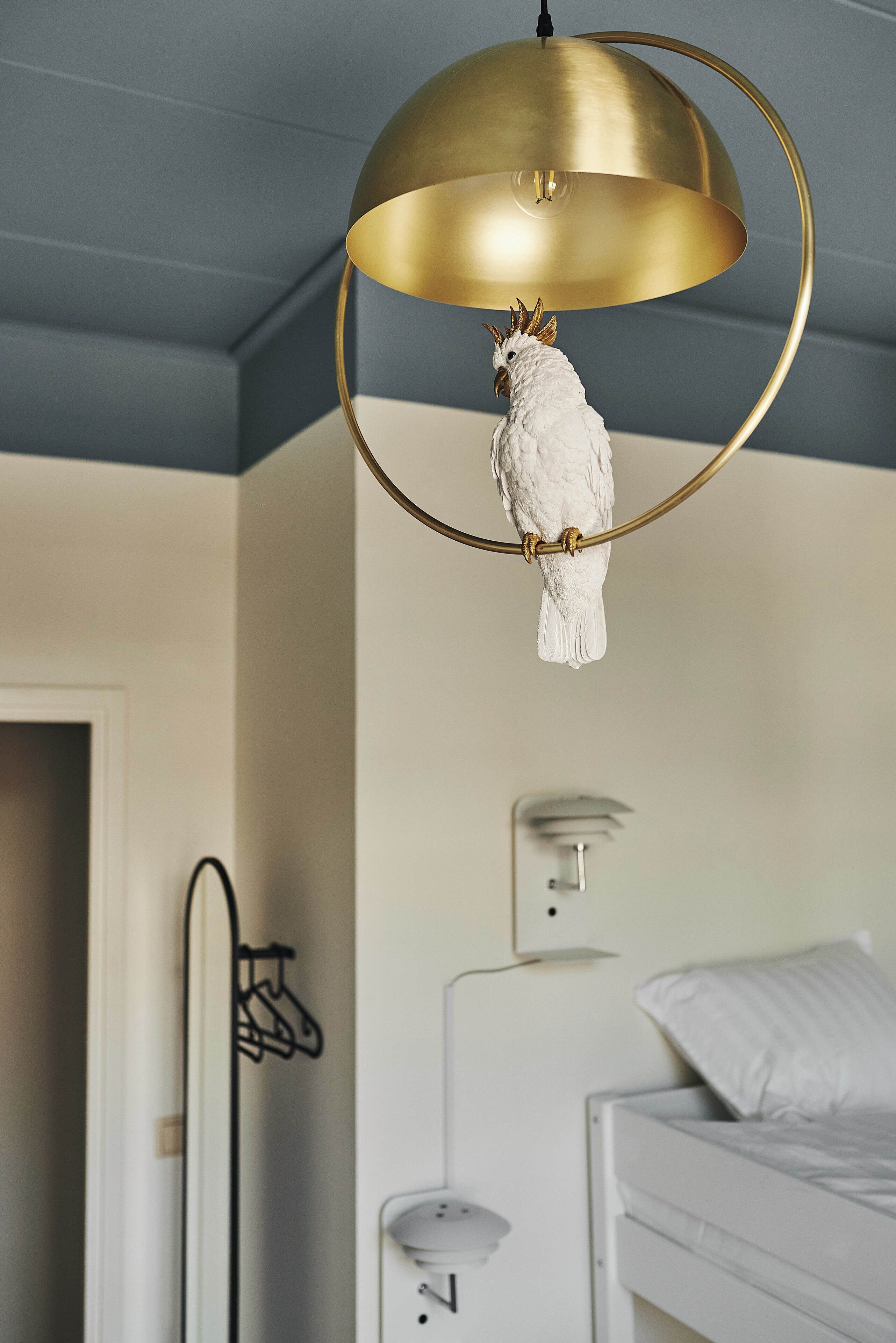 Un ave blanca posada sobre una lámpara con aro dorado en una habitación con cama y pared blanca.