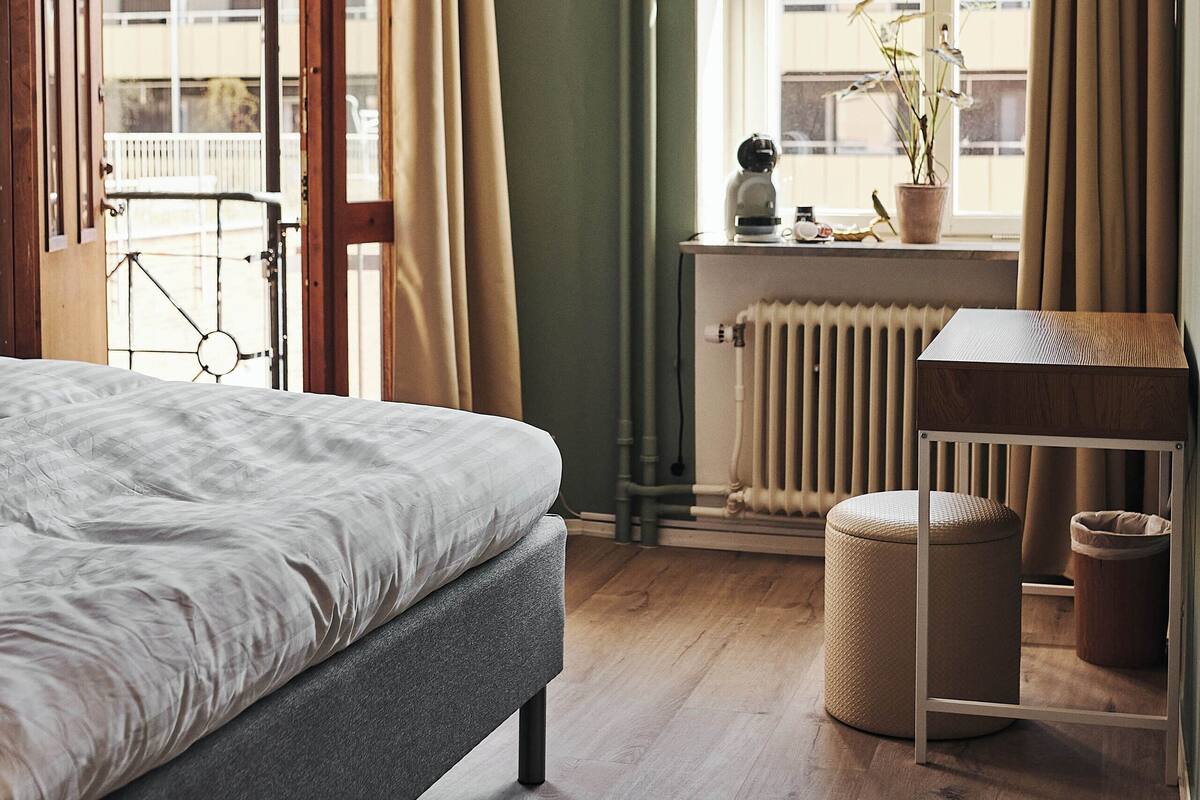 Un dormitorio con cama, escritorio, radiador, ventana con cortinas y vista a edificios.