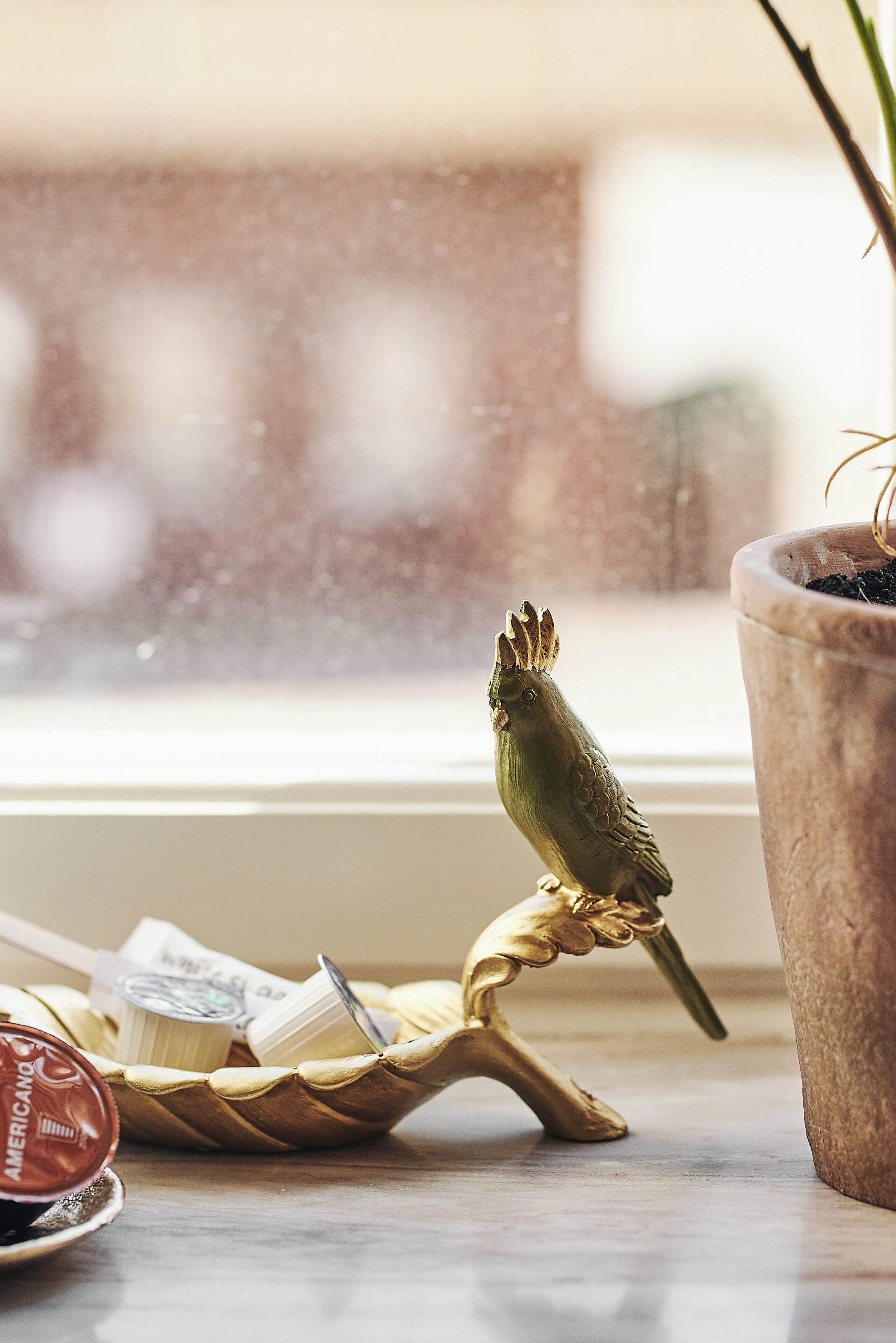 A bird figurine on a decorative stand next to a window with a blurred view outside.