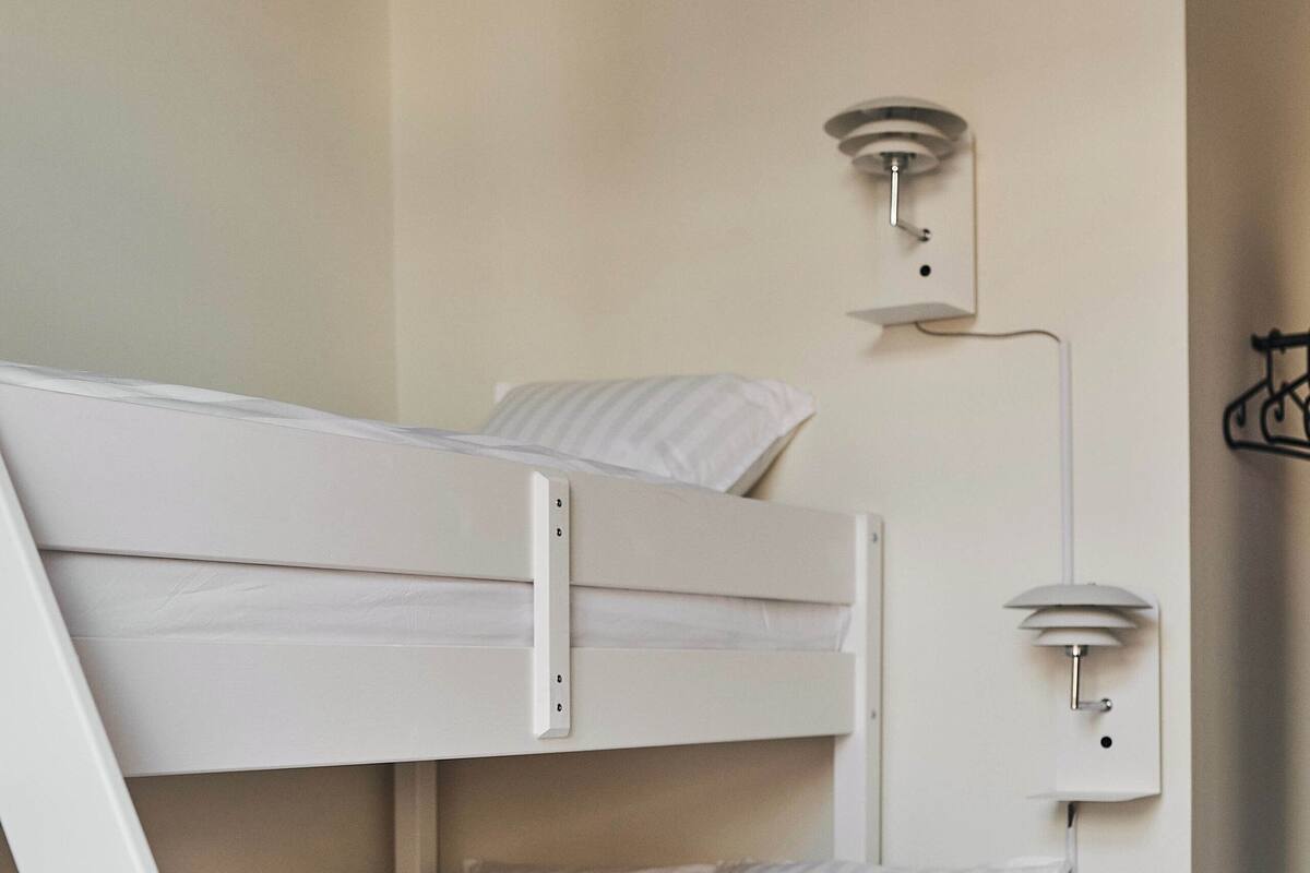 Una cama litera con estructura y ropa de cama blancos, un aplique en la pared y un perchero.