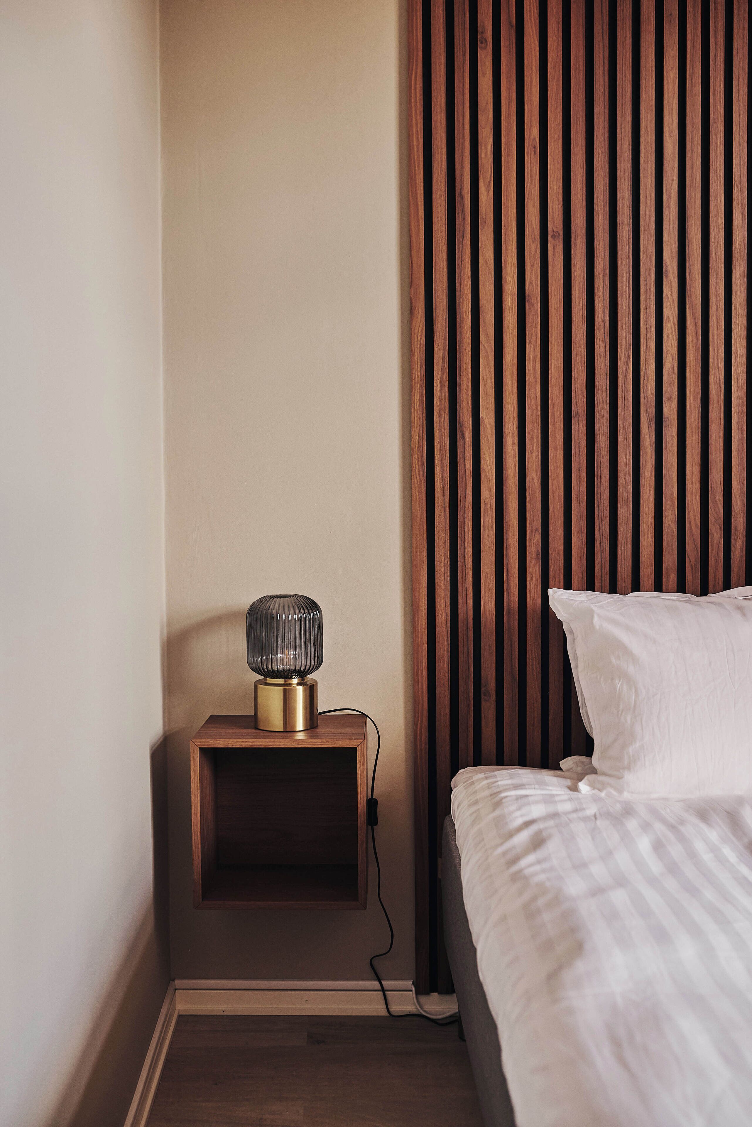 A neatly made bed with white linens, a bedside table with a lamp, and a wooden slat headboard.