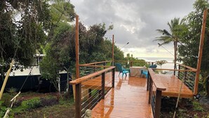A wooden deck with chairs and a statue, surrounded by plants and a view of the sea. - Manta Suite w/ ocean view and sleeping 6 --TA-154-746-2656-01 (Kealakekua)