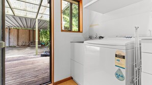 A compact, modern bathroom with a large washing machine, a window with a wooden frame, and a view of a wooden deck and greenery outside. - The Wee Bothy - Onemana Holiday Home (Onemana)