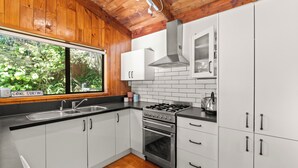 A modern kitchen with white cabinets, a stainless steel stove, and a window overlooking greenery. - The Wee Bothy - Onemana Holiday Home (Onemana)