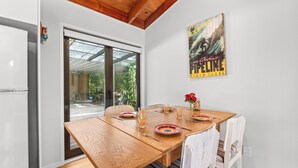 A dining area with a wooden table, chairs, and a glass door leading to a garden. - The Wee Bothy - Onemana Holiday Home (Onemana)