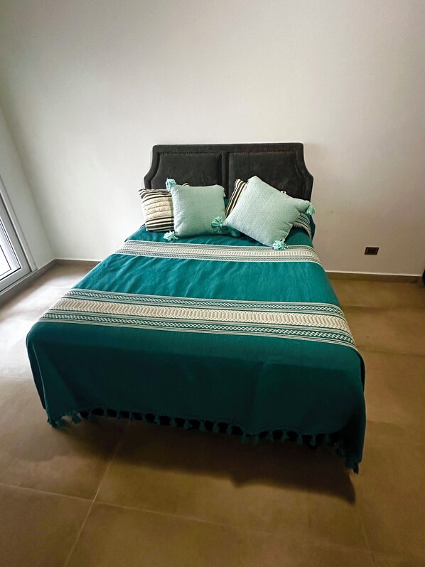 A bed with a teal bedspread and decorative pillows, a dark headboard, and a plain wall.