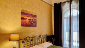 A hotel room with a bed, a painting of a sunset scene, a lamp, and a window with curtains.