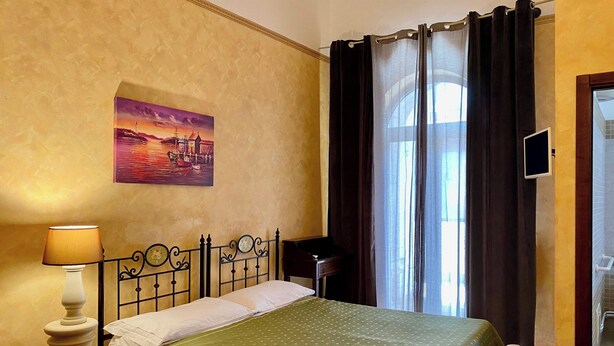 A hotel room with a bed, a nightstand, a lamp, a painting on the wall, and a view of the outside through the window.