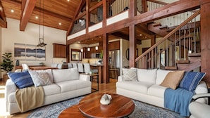 Ein großzügiger Wohnbereich mit zwei weißen Sofas, einem Holztisch und einem großen Teppich. Eine Treppe mit Holzhandläufen führt in eine obere Ebene.