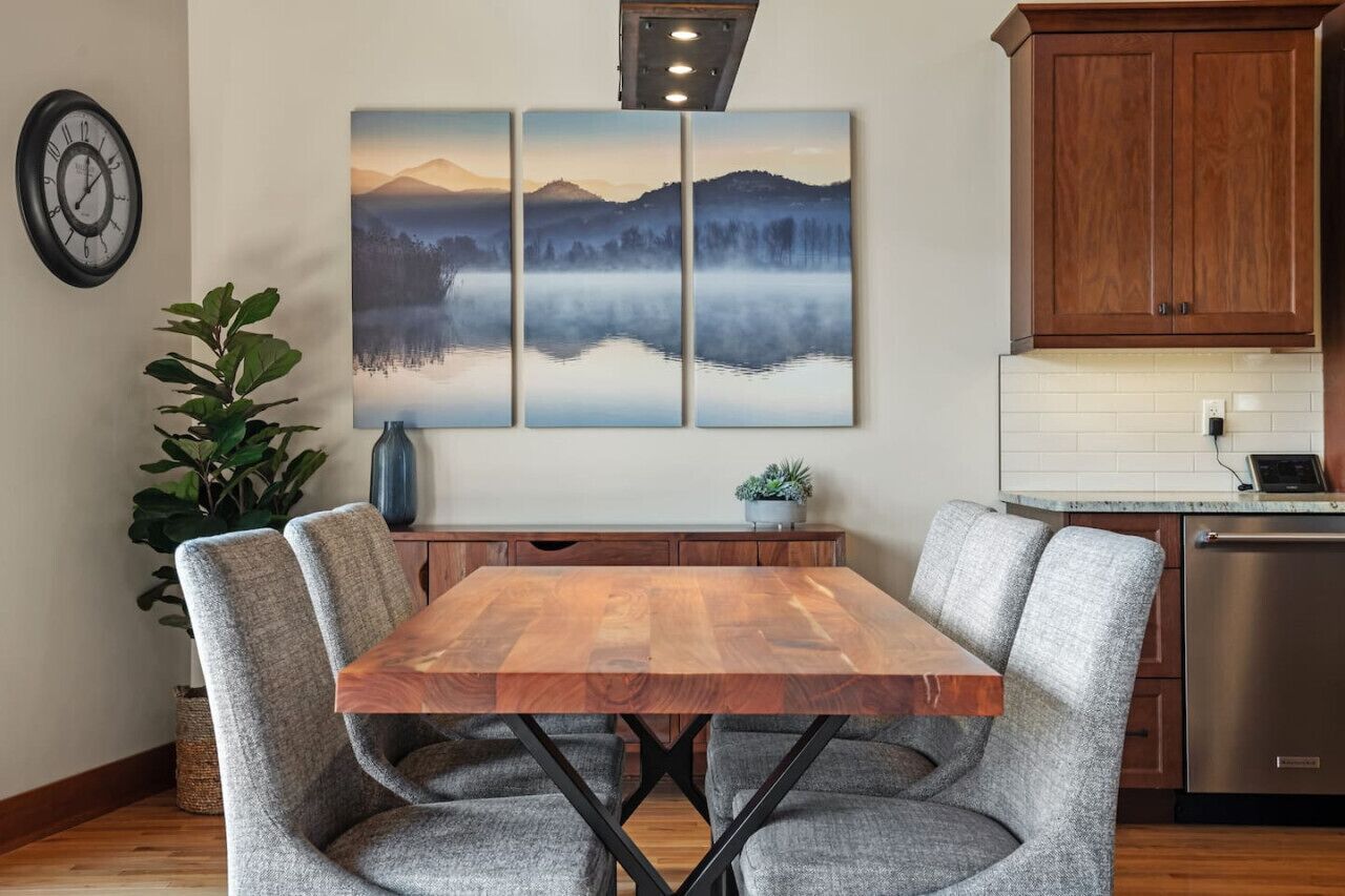 A dining area with a wooden table and grey upholstered chairs, a large landscape painting above, and a kitchen with wooden cabinets and a stainless steel dishwasher.