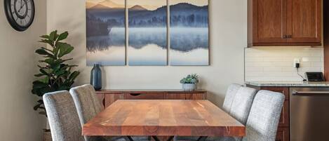 A dining area with a wooden table and grey upholstered chairs, a large landscape painting above, and a kitchen with wooden cabinets and a stainless steel dishwasher.