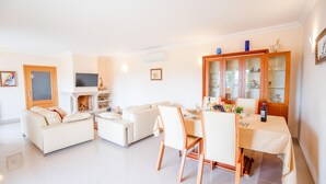 Un salon moderne avec une cheminée, des canapés blancs, une table à manger dressée pour quatre personnes et un buffet en bois doté de portes en verre.