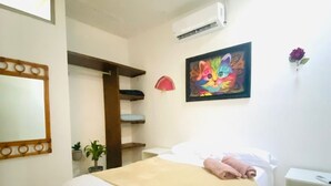 A bedroom with a bed, a ceiling fan, a painting, an air conditioner, and a small table.