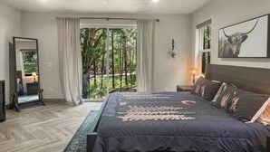 Ein modernes Schlafzimmer mit einem großen Bett, einem Nachttisch, einem Spiegel und Blick auf einen Wald durch eine Schiebetür.