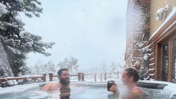 Ett bubbelbad för två personer, omgivet av snötäckta träd och en byggnad av sten.