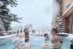 A hot tub with two people, surrounded by snow-covered trees and a stone building.