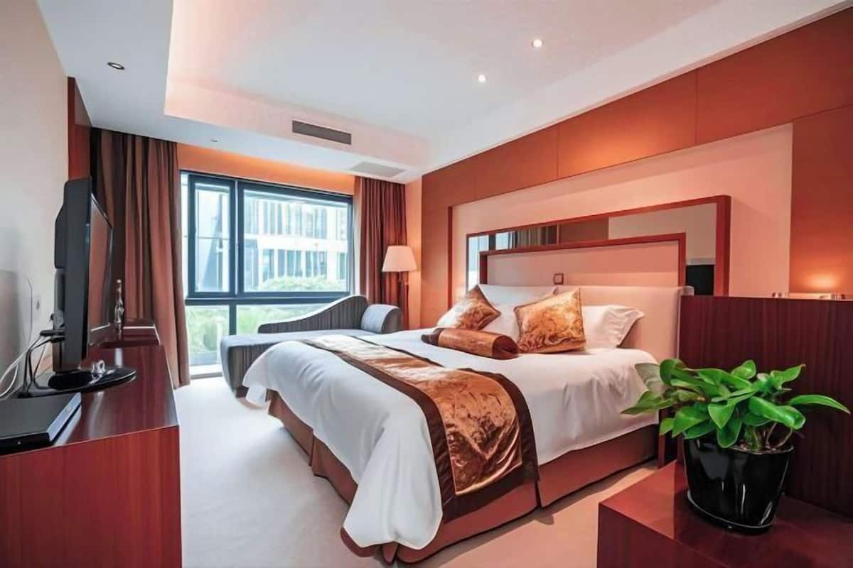 A modern hotel room with a large bed, a desk, a television, and a window with a view of buildings.