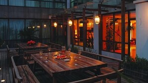 Un espacio de comedor al aire libre con mesas y bancos de madera, iluminación ambiental y vistas del interior a través de puertas de cristal.