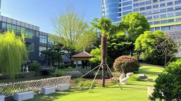 A landscaped garden with a gazebo, a palm tree, and a pond, surrounded by modern buildings.