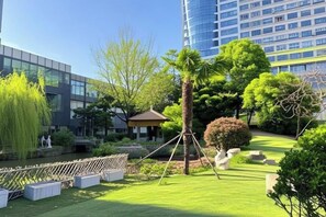 Un jardín ajardinado con un pabellón, una palmera y un estanque, rodeado de edificios modernos.