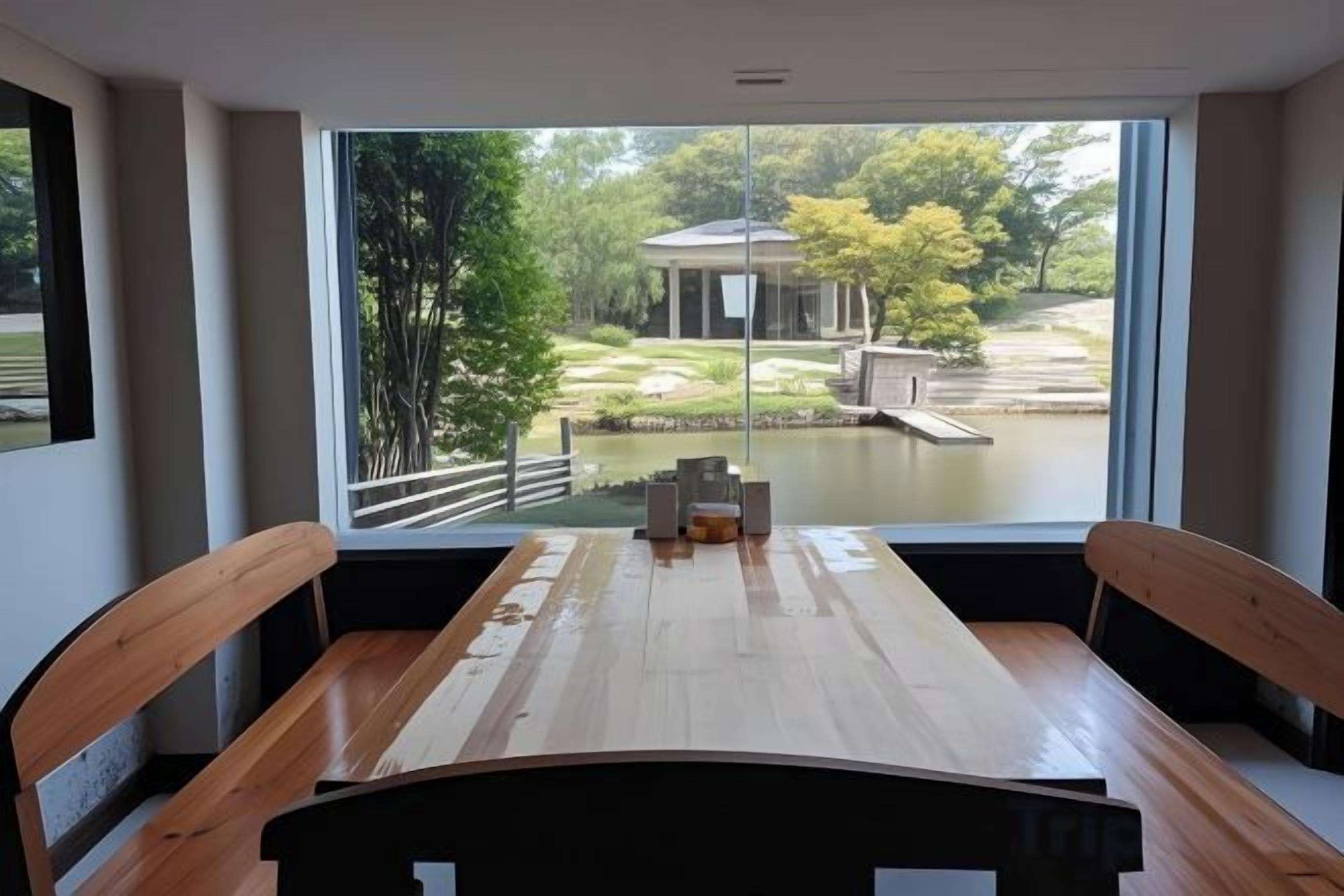A dining area with a wooden table and chairs, a view of a garden and pond through large windows.
