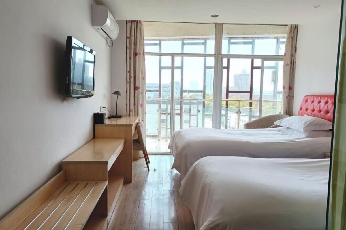 A hotel room with two beds, a TV mounted on the wall, a wooden bench, and a balcony with a view of the city.
