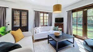 Ein modernes Wohnzimmer mit einem weißen Sofa, einem dunklen Couchtisch und einem Fernseher. Es gibt ein großes Fenster mit Vorhängen, eine Vitrine und eine Pendelleuchte.