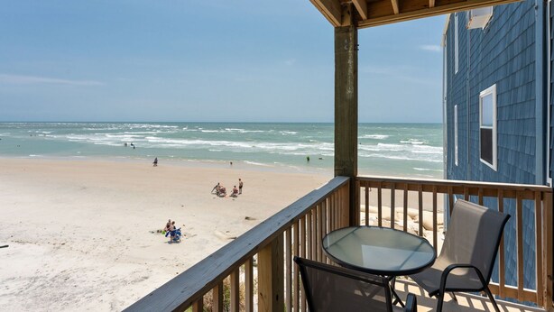 Une terrasse en bois avec une table et des chaises, donnant sur une plage où l’on peut voir des gens et des vagues.