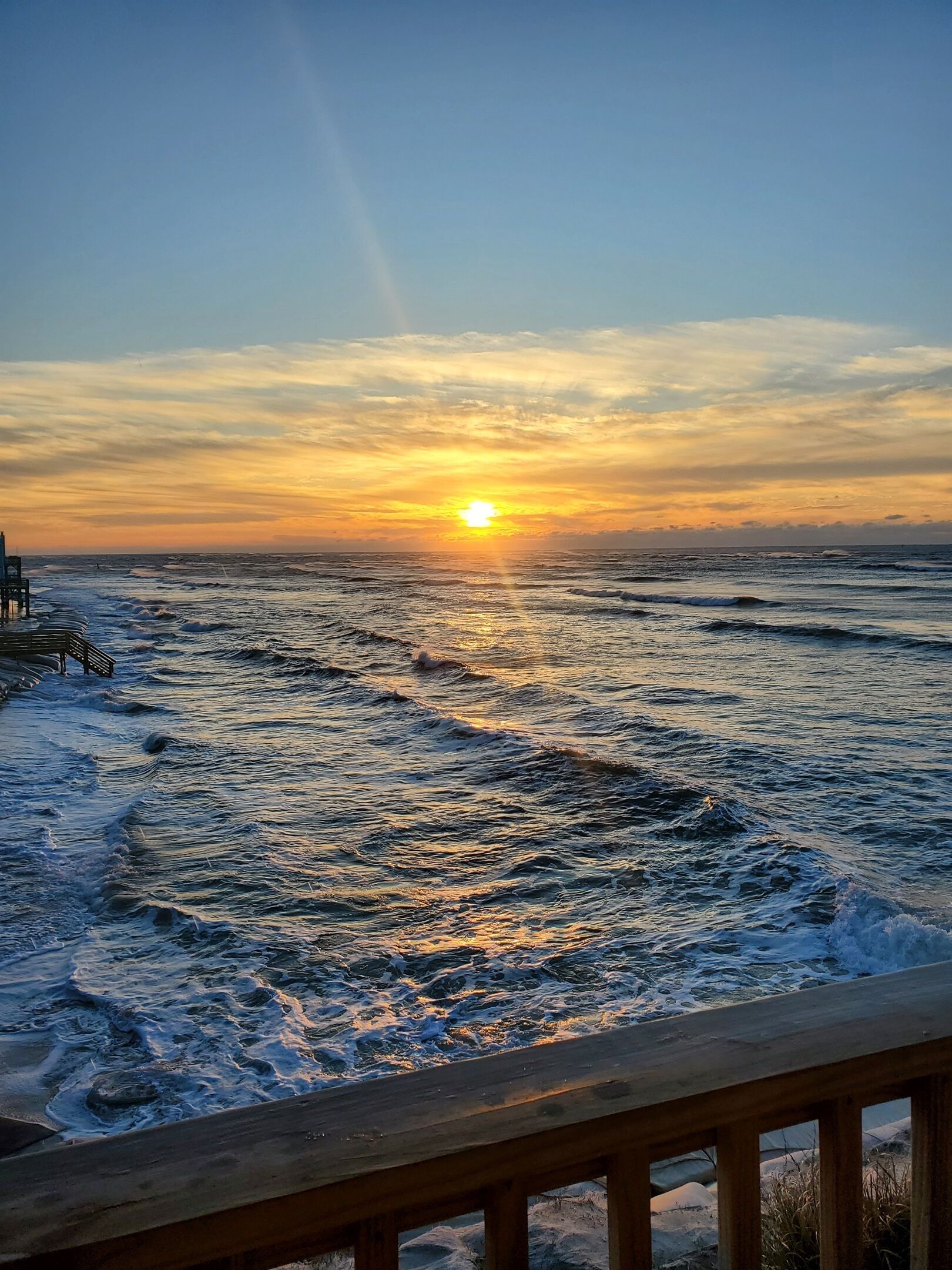 A wooden deck overlooking a beach with waves and a sunset.