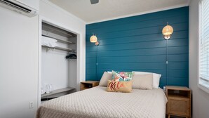 Une chambre à coucher avec une tête de lit de couleur turquoise, un lit recouvert d’une courtepointe beige, deux lampes de chevet, une table de chevet et une armoire où sont suspendus des vêtements.