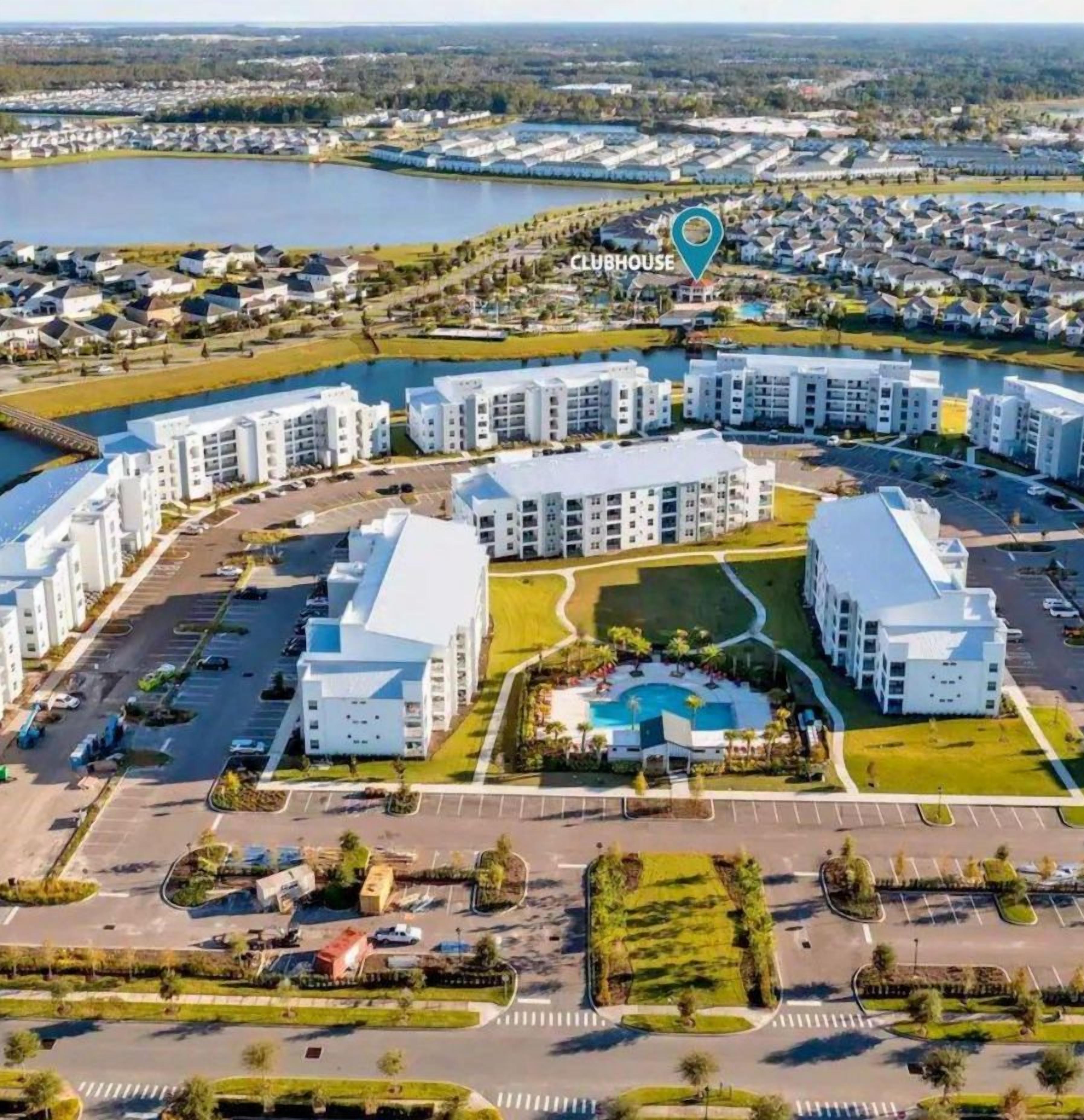An aerial view of a modern residential complex with multiple buildings, a central swimming pool, and a surrounding green space.