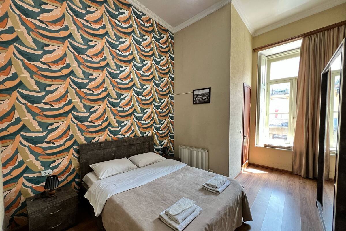 A bedroom with a patterned wallpaper, a bed with white linens, a bedside table with a lamp, and a window with curtains.