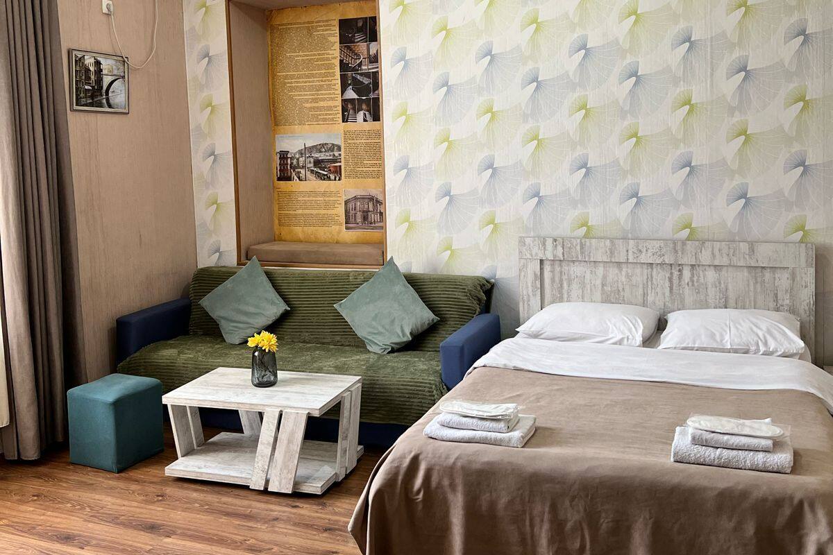 A hotel room with a bed, a sofa, a small table, and a wall-mounted air conditioner.