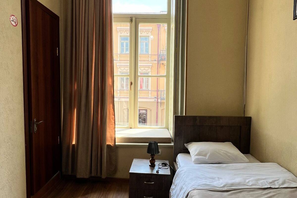 A hotel room with a wooden floor, a bed with white linens, a nightstand with a lamp, a window with curtains, and an air conditioning unit.