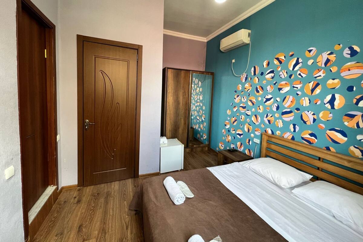 A bedroom with a wooden bed, a white bedside table, a wall with a colorful abstract design, and a ceiling light.