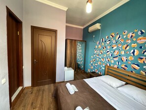 A bedroom with a wooden bed, a white bedside table, a wall with a colorful abstract design, and a ceiling light. - Hotel Royal Bridge (Tbilisi)