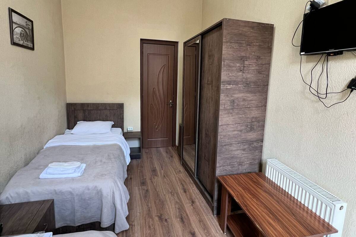 A hotel room with two single beds, a wooden wardrobe, a small table, and a TV mounted on the wall.