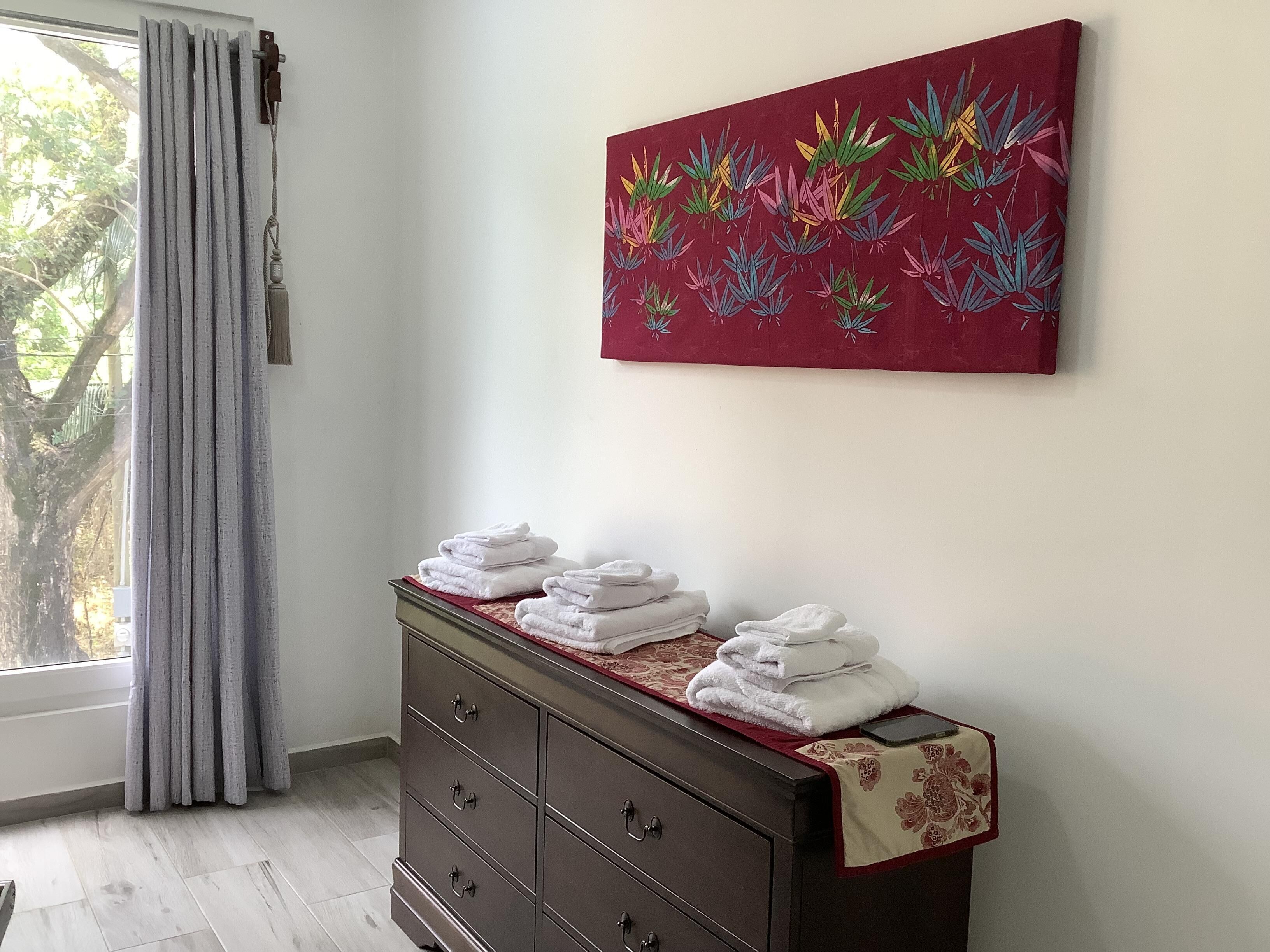 Ein Zimmer mit einem Holzkasten, einem Fenster mit Vorhängen und einem farbenfrohen Bild an der Wand.