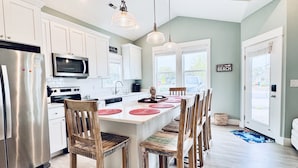 Una cocina moderna con armarios blancos, un refrigerador de acero inoxidable, un microondas y una isla grande con individuales rojos. Hay sillas de madera alrededor de la isla y una lámpara colgante arriba.