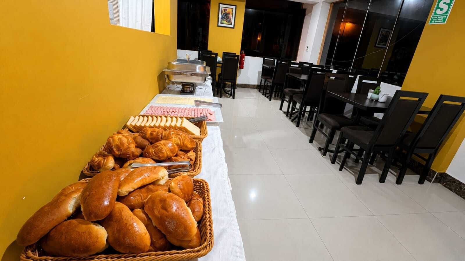 Mesa de buffet con pan y repostería, mesas y sillas dispuestas para comer.