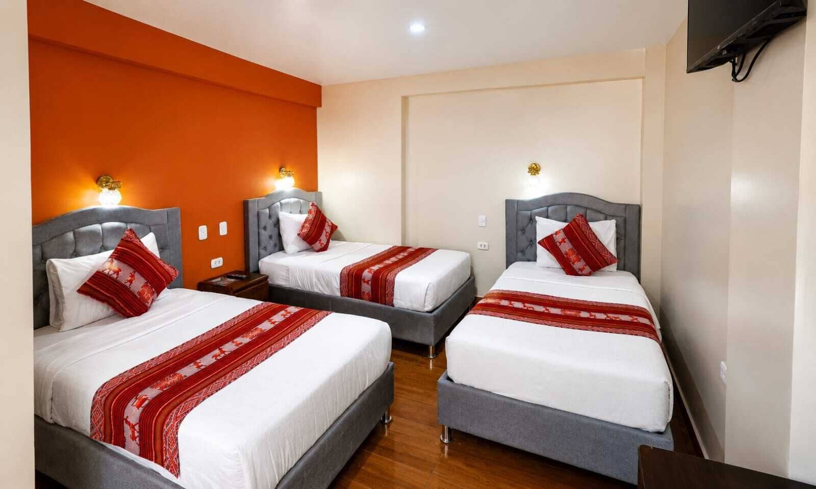 Habitación de hotel con tres camas, cada una con cabecera gris y ropa de cama blanca, y almohadas decorativas rojas.