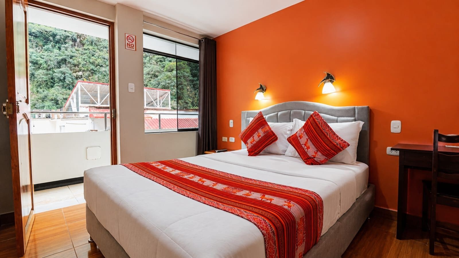 A hotel room with a bed, a desk, and a view of a red-roofed building through a window.