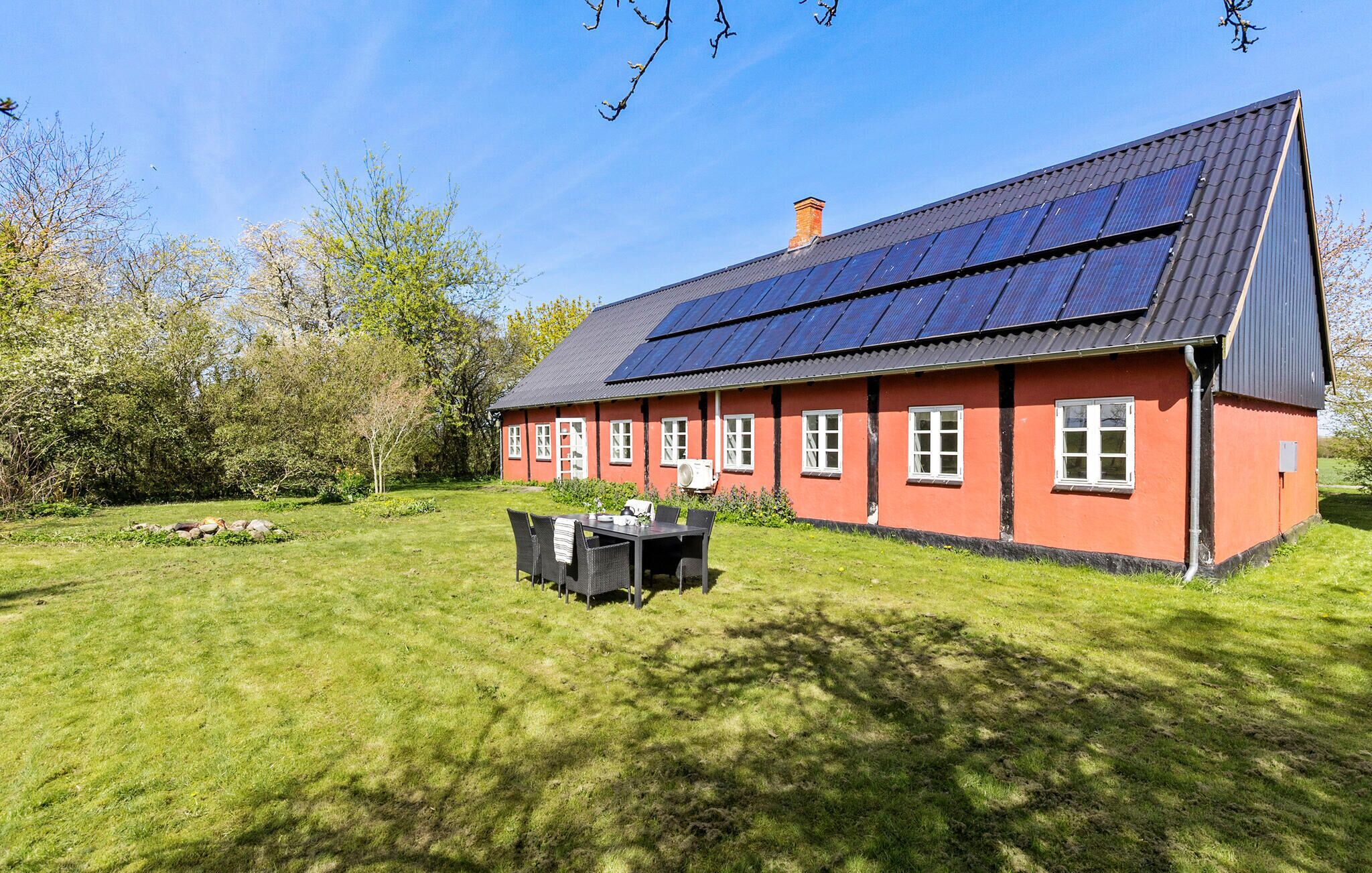 A red house with solar panels on the roof, a lawn, and a table with chairs in front.