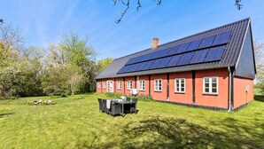 A red house with solar panels on the roof, a lawn, and a table with chairs in front.