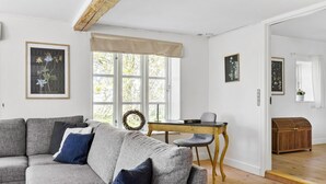A living room with a grey sofa, a small wooden table, and a wicker chair. There are framed botanical prints on the walls and a wooden chest of drawers in the corner.