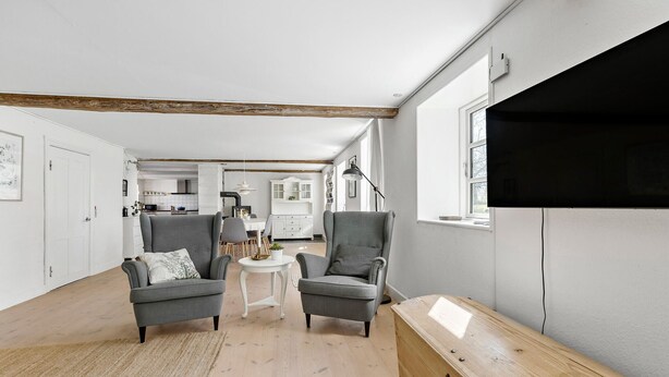 A modern living room with two grey armchairs, a small white table, and a wooden bench. A kitchen and dining area are visible in the background.